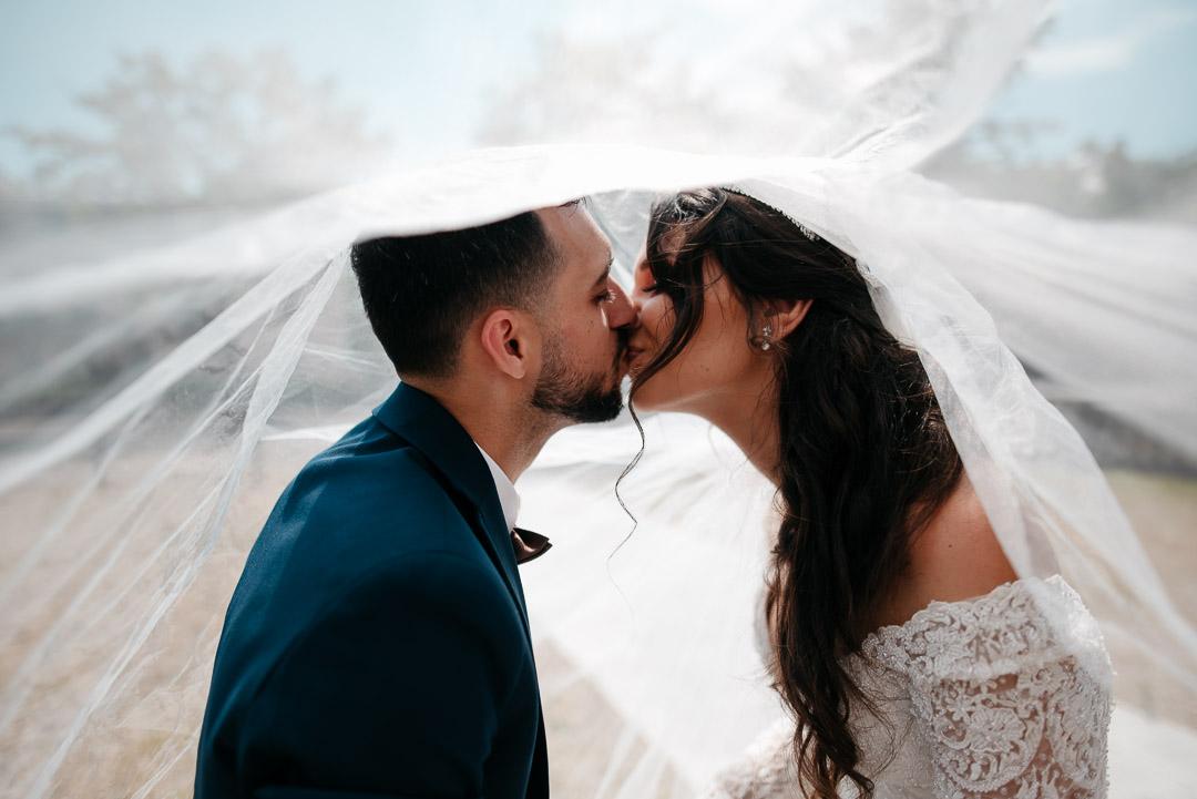 Couple de mariés s'embrassant sous le voile blanc lors d'une séance photo de mariage en extérieur en Alsace.